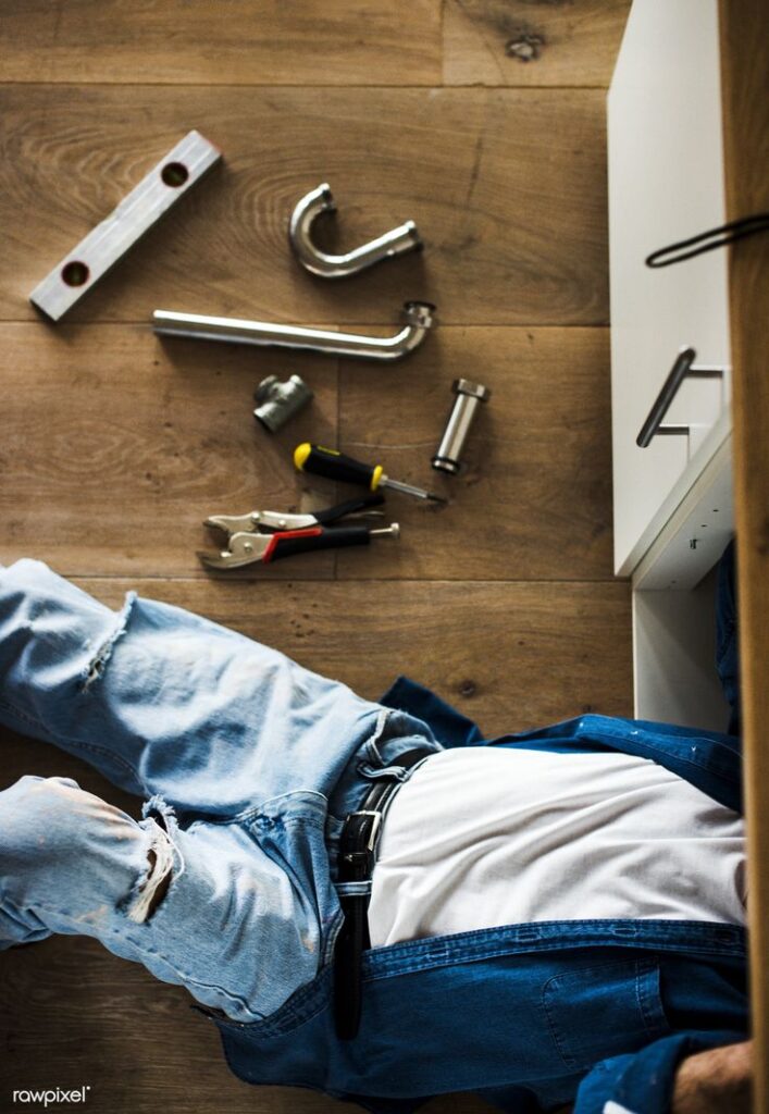 Plumber fixing pipes under sink, tools scattered on wooden floor, jeans and tool belt visible.