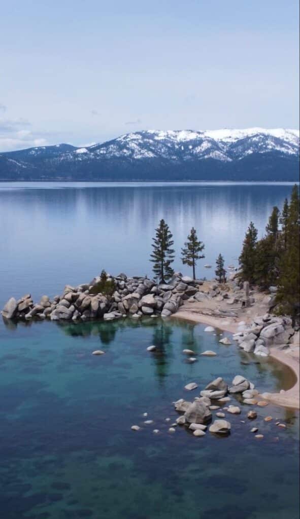 Tranquil lake scene with clear water, rocky shoreline, evergreen trees, and snow-capped mountains in the background.