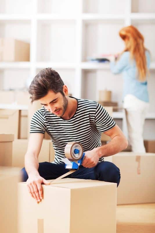 Man packing cardboard boxes with tape gun for moving, while a woman organizes in the background.