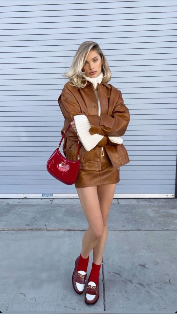 Elsa Hosk winter outfit_ Helsa Studio sweater Woman in brown leather outfit with red handbag stands against a gray backdrop, styled with red socks and loafers. | Sky Rye Design Woman in brown leather outfit with red handbag stands against a gray backdrop, styled with red socks and loafers.