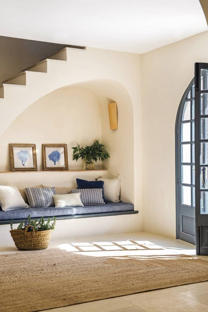 Esta hermosa masía en Mallorca estaba abandonada y Cozy nook with blue bench, decorative pillows, framed art, and woven basket under staircase. Sunlight through arched door. | Sky Rye Design Cozy nook with blue bench, decorative pillows, framed art, and woven basket under staircase. Sunlight through arched door.