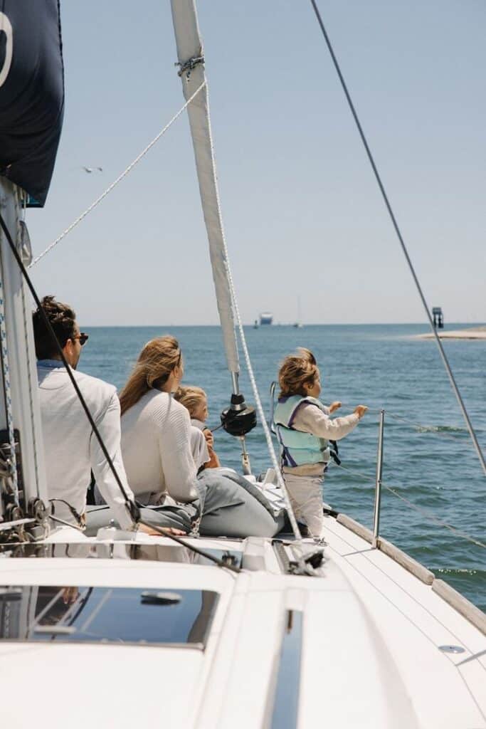 Family enjoying sunny day on a sailboat, sailing on calm ocean waters under a clear blue sky.