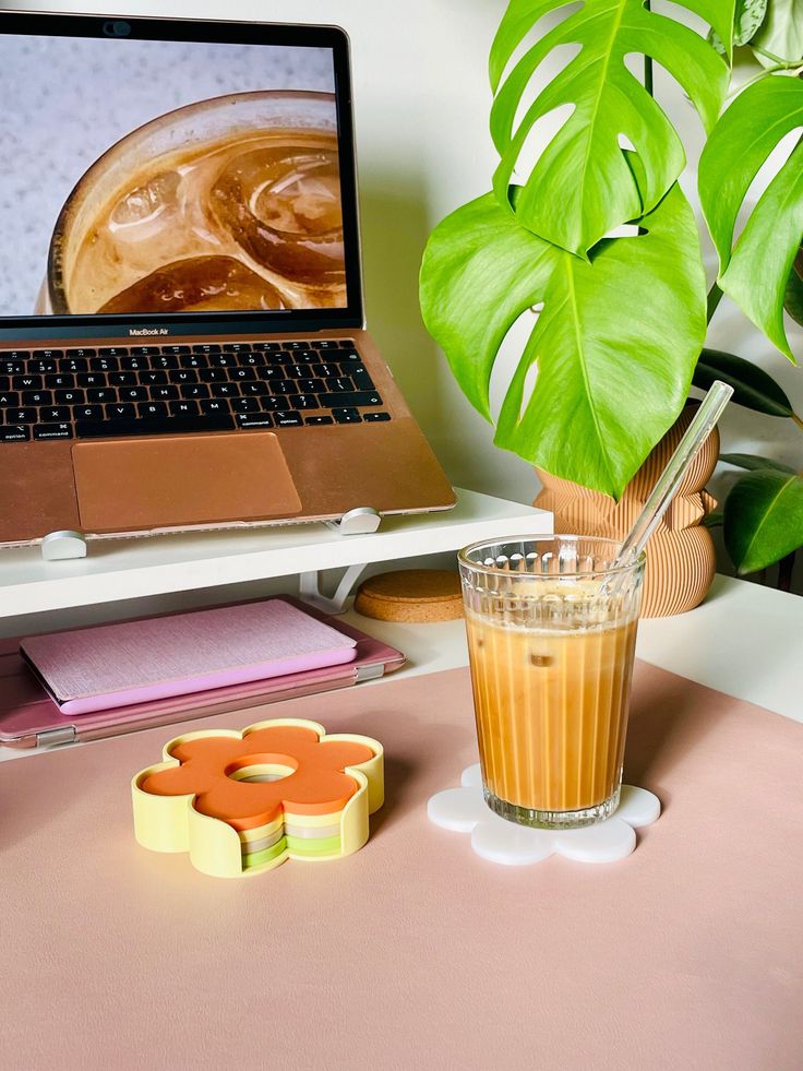 Cozy workspace with iced coffee, laptop, plant, and colorful coasters on a desk, perfect for productive mornings.