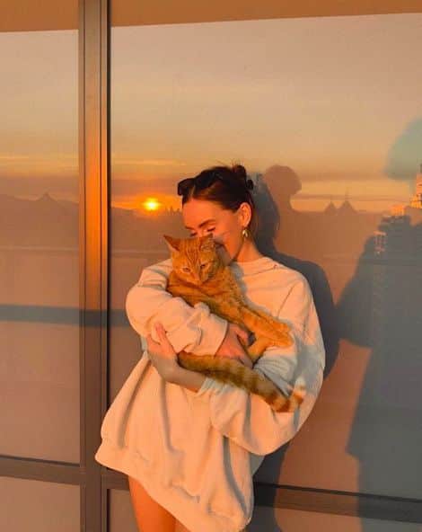 Woman enjoys sunset on balcony, holding a ginger cat, reflecting warm hues on glass door.