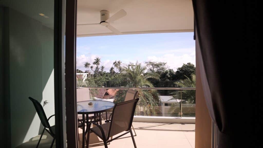 Balcony view with chairs and table overlooking palm trees and rooftops under a blue sky.