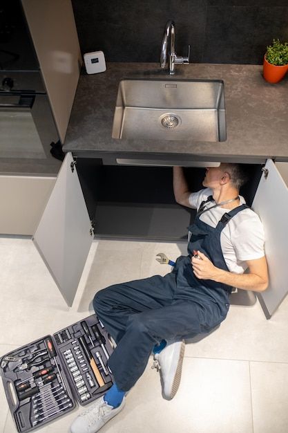 Plumber fixing kitchen sink, lying under the cabinet with a toolbox nearby, wearing blue overalls and a white shirt.