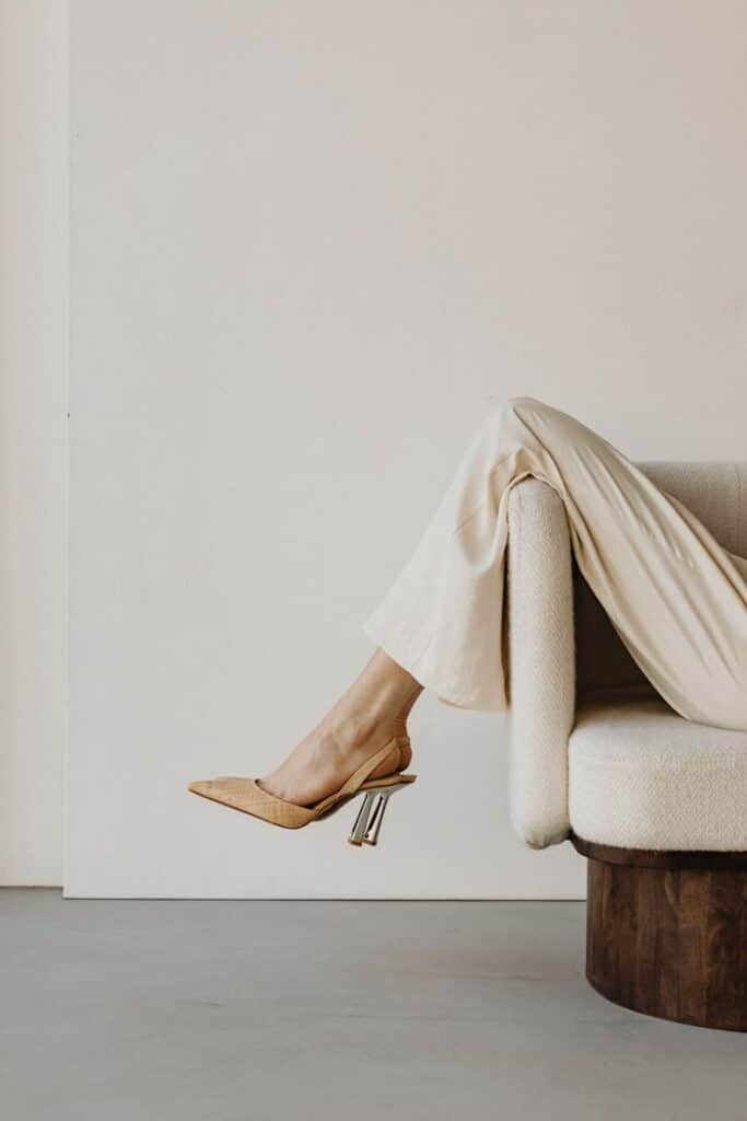 Beige stiletto heels on relaxed legs draped over a cream sofa, minimalist modern style.