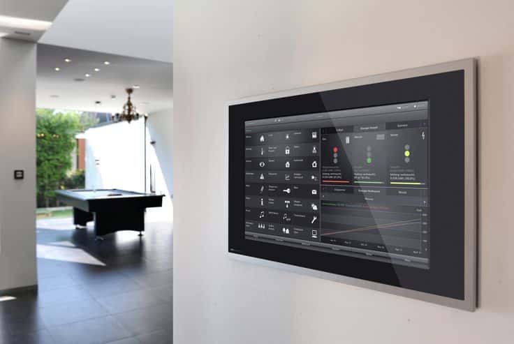 Smart home control panel on a wall with modern interior in the background, featuring a pool table and chandelier.