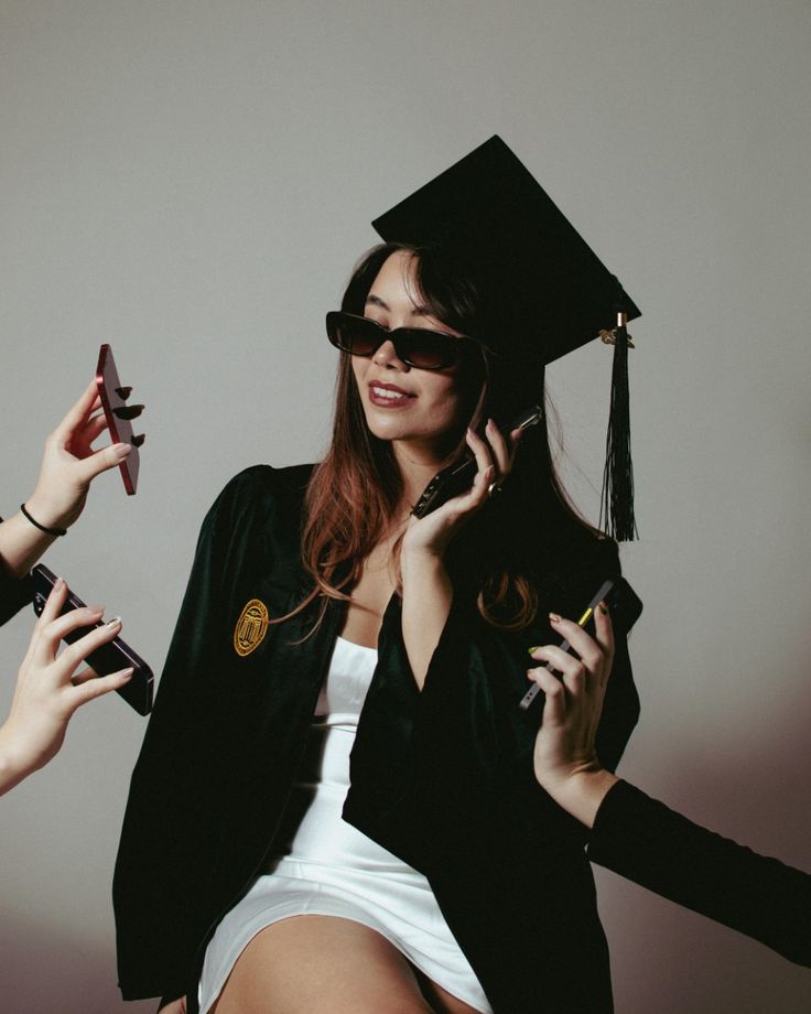 Graduate wearing cap and gown, multitasking with phones, smiling confidently in sunglasses.