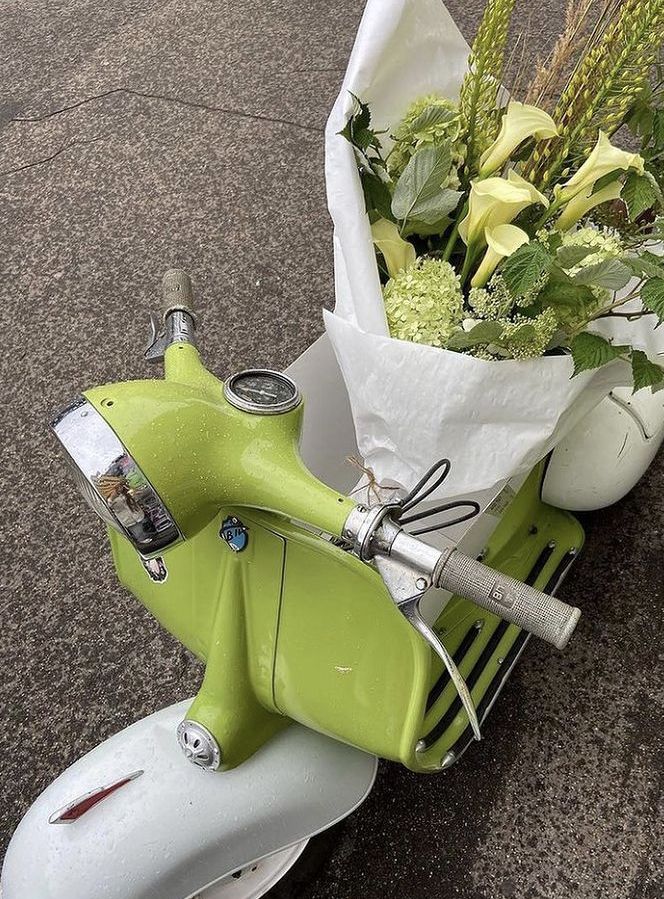 Vintage green scooter with a white basket holding fresh lilies and greenery on a wet asphalt background.