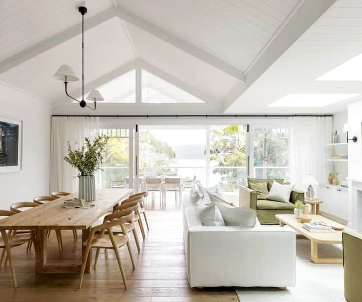 Bright, airy living-dining room with wooden table, white sofa, and a view of greenery through large windows.