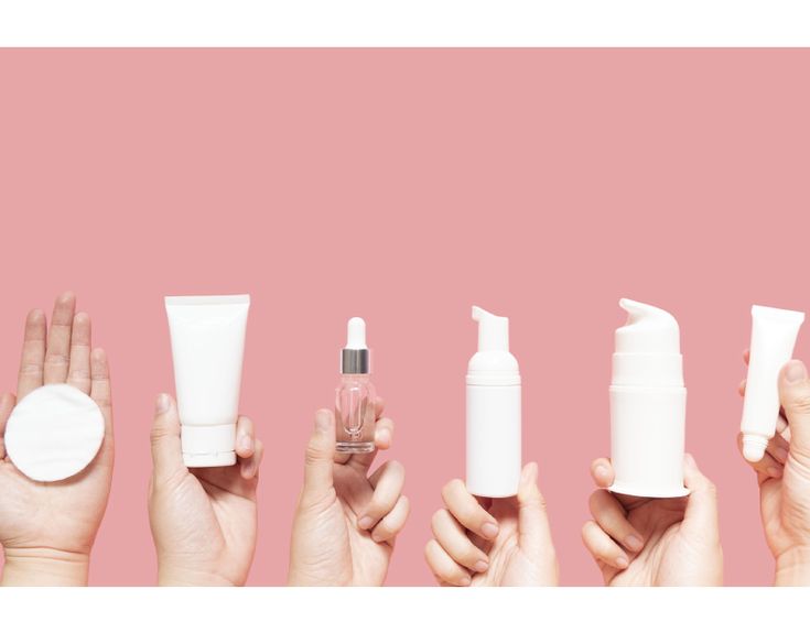 Hands holding skincare products including lotion, serum, and foam dispenser against a pink background.