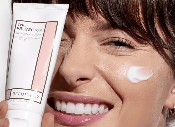Woman smiling with sunscreen on face, holding The Protector SPF 30 cream tube close-up.