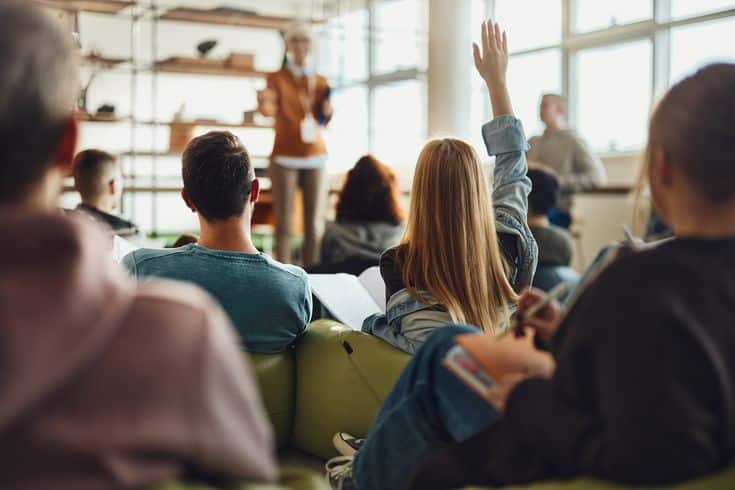 Students in a classroom, one raising hand, engaging with teacher. Bright, modern learning environment with natural light.