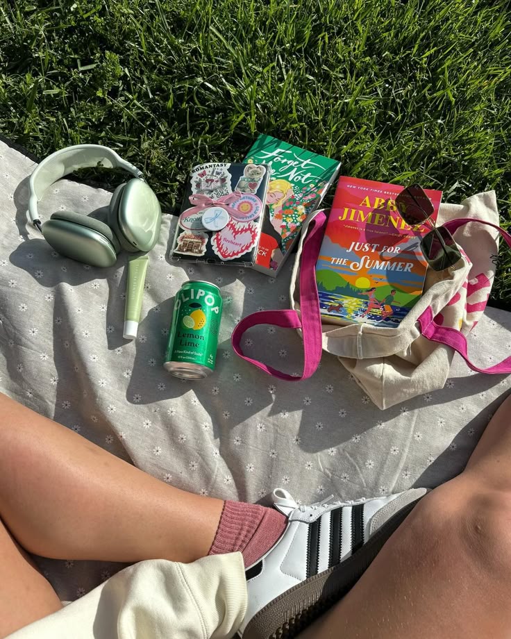 Outdoor picnic scene with books, headphones, a soda can, and sneakers on a blanket in the grass.