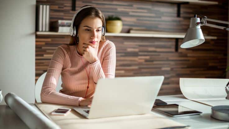 Woman focused on laptop, wearing headphones at a desk with a tablet and phone, engaging in remote work or study.