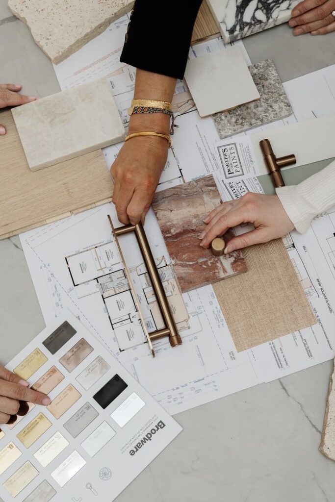 Hands selecting building materials and fixtures over blueprints, featuring stones, fabrics, and metal finishes.