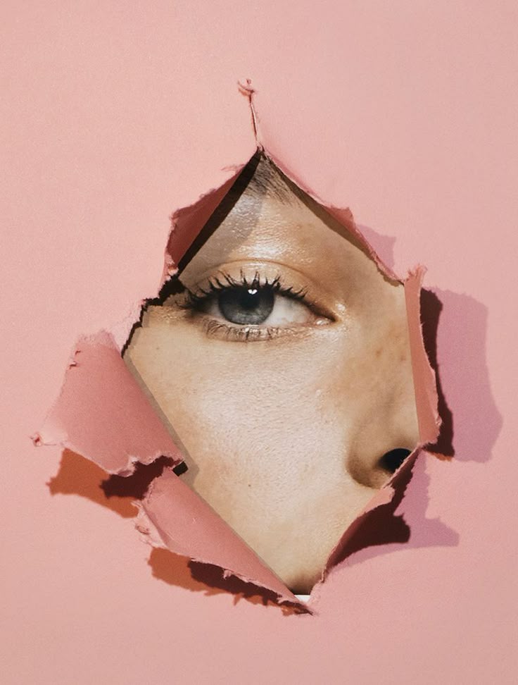 Close-up of a blue eye looking through a ripped pink paper, symbolizing curiosity and exploration.