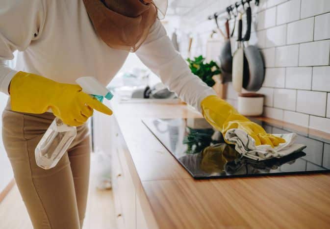 Person cleaning kitchen stove with spray and cloth, wearing yellow gloves for hygiene.