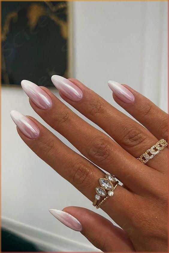 Elegant hand with pearlescent white-pink almond nails and gold rings, showcasing a stylish manicure.
