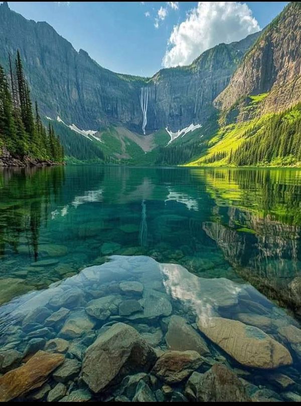 Crystal-clear lake reflecting mountains and waterfall under a blue sky, surrounded by lush greenery and rocky shores.