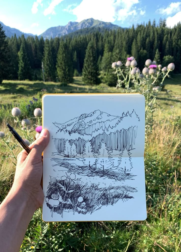 Sketch of a mountain landscape with trees and flowers, held against the real scenery in sunlight.