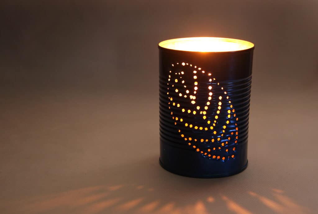 Homemade tin can lantern with cutout leaf design, glowing warmly in dim light.