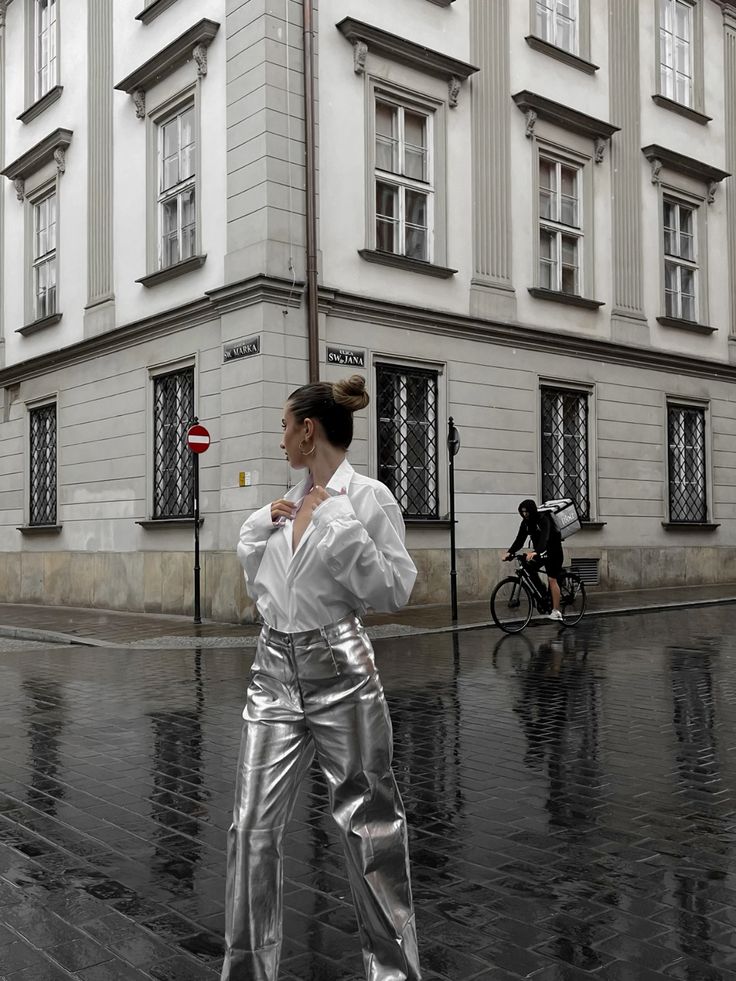 Fashionable woman in metallic pants poses on a rainy city street with a cyclist in the background.