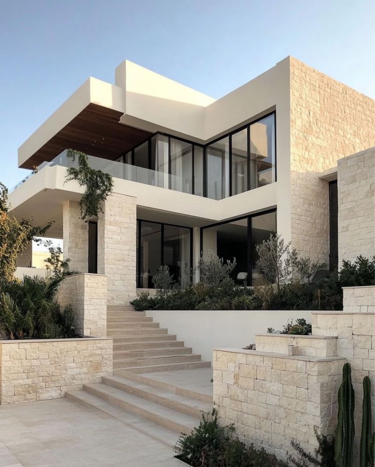 Modern luxury home with large windows and stone facade, featuring lush greenery and a sky backdrop.