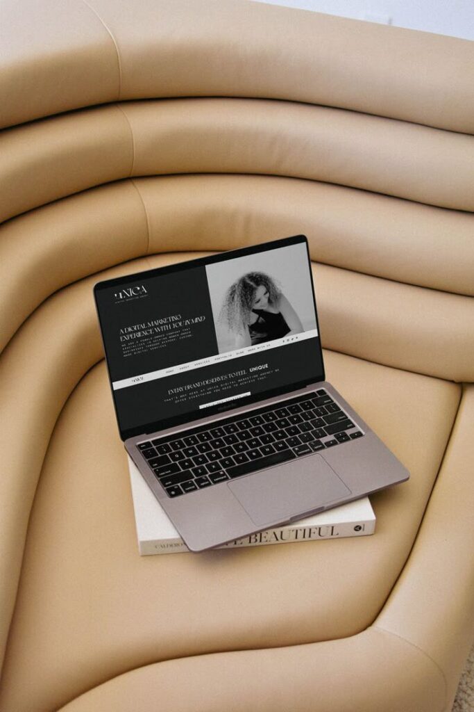 Laptop displaying a website on a beige sofa with books underneath, creating a cozy, modern workspace.