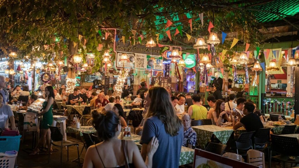 Bustling outdoor restaurant with colorful lights and decor, filled with people dining and socializing under leafy canopy.