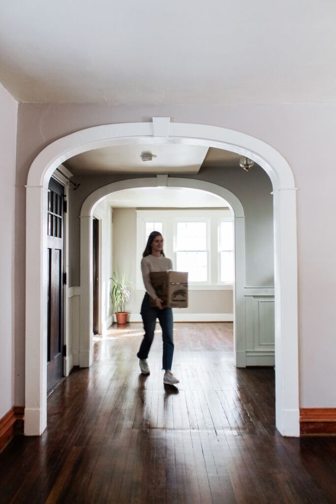 Person carrying box in a bright, modern hallway with arched entry and wooden floors.