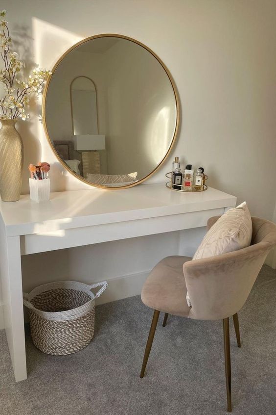 Elegant vanity setup with round mirror, cosmetics, and decor on a white desk and beige chair in a bright room.