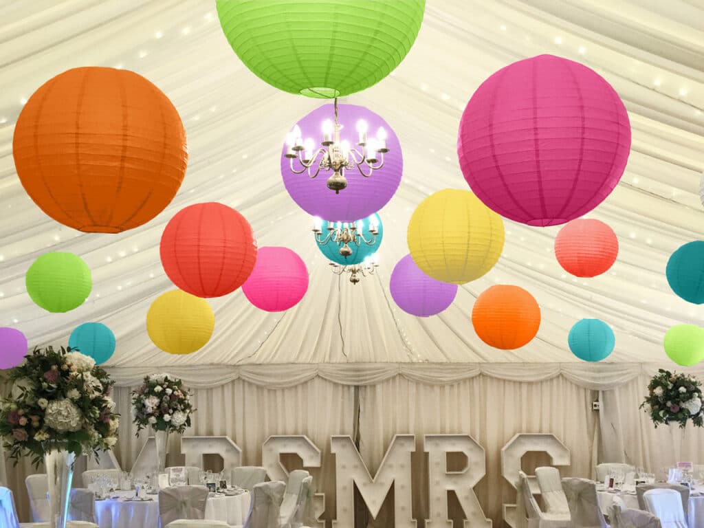 Colorful paper lanterns and floral centerpieces decorate an elegant tented wedding reception with illuminated chandeliers.