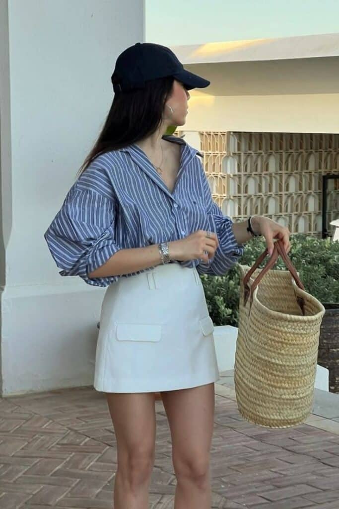 Must-Have Beach Outfits for Summer Tap to Shop Now Woman in striped shirt and skirt holding woven bag, wearing a cap, looking stylish outdoors. | Sky Rye Design Woman in striped shirt and skirt holding woven bag, wearing a cap, looking stylish outdoors.