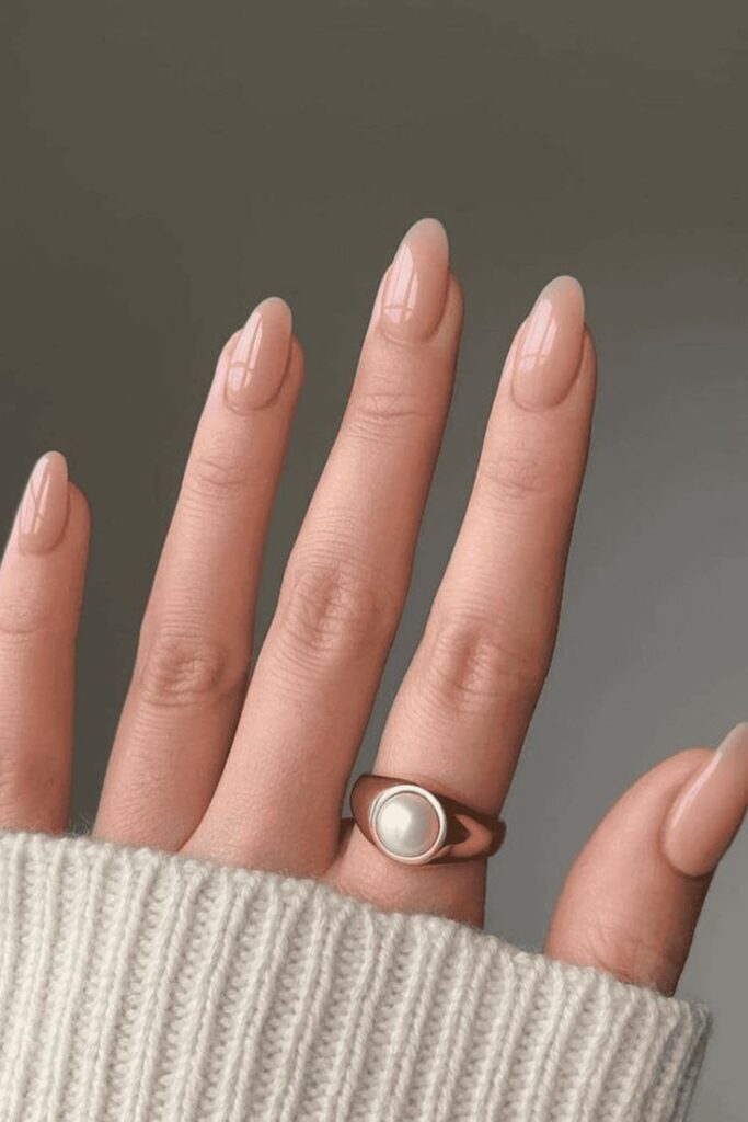 Close-up of a hand with nude almond nails, wearing a pearl ring and gray sweater, perfect for a minimalist look.