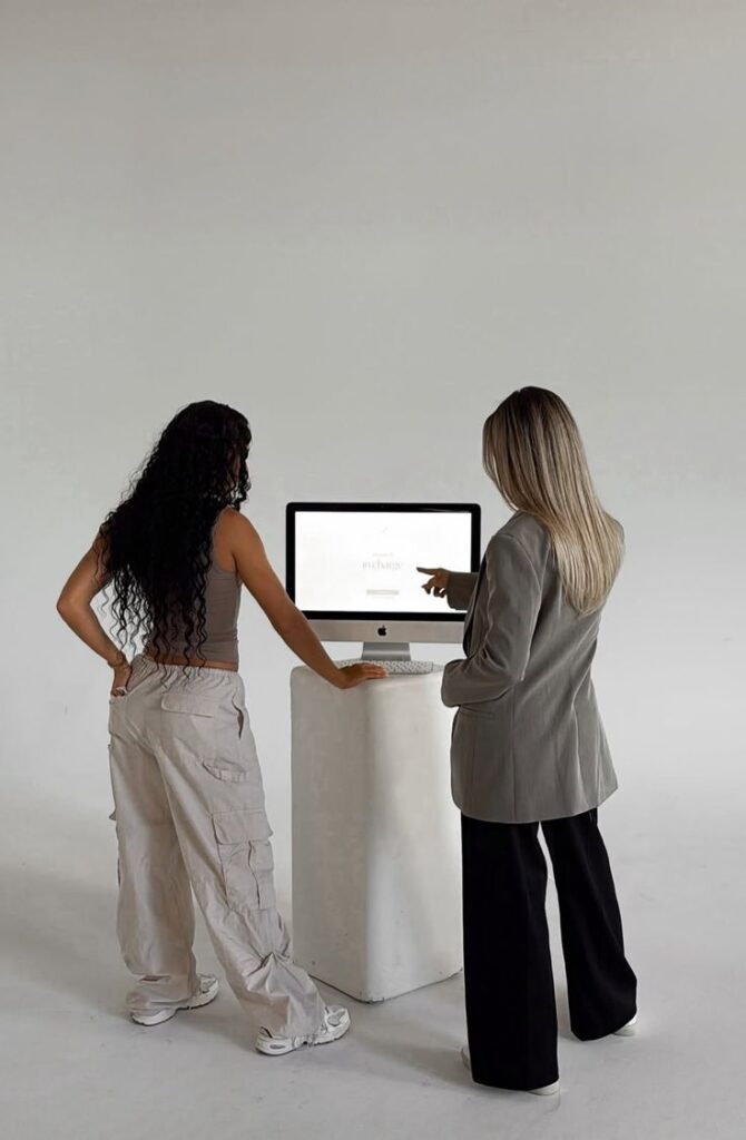 OLIVIA BRAND Branditbabe _ Stan Two women collaborate in front of a desktop computer in a minimalist setting. | Sky Rye Design Two women collaborate in front of a desktop computer in a minimalist setting.