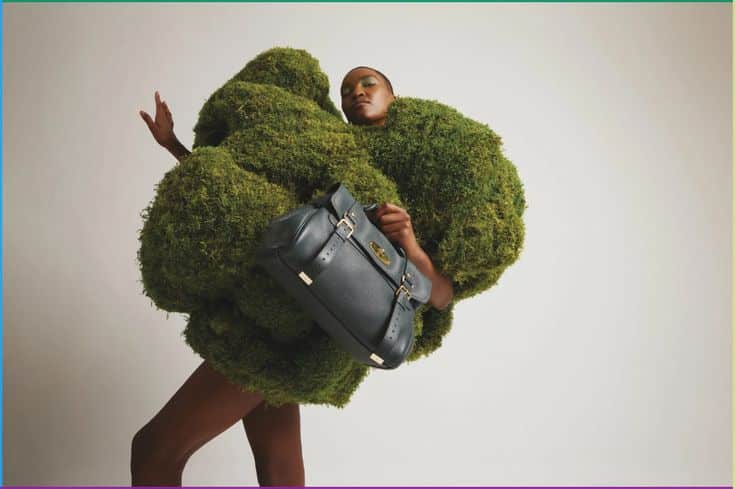 Model in a moss-like outfit poses with a black designer handbag, showcasing avant-garde fashion style.