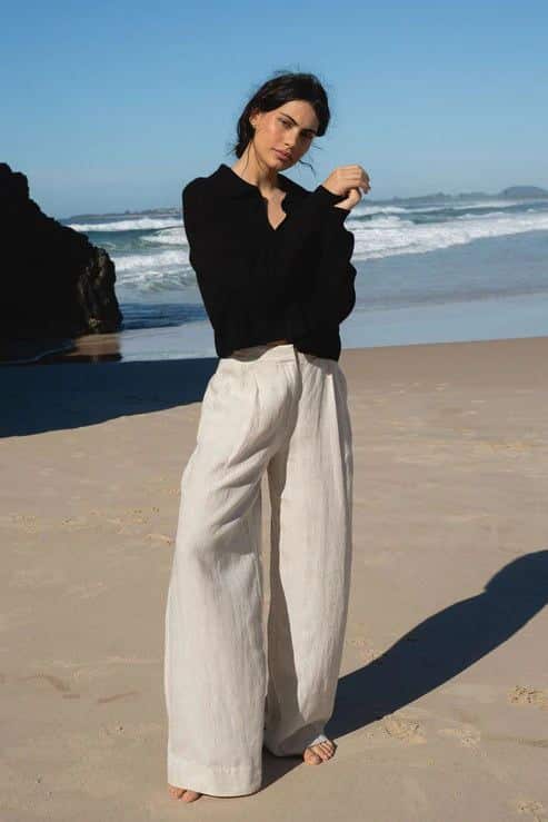 Woman in black top and wide beige pants stands on a sunny beach with a bright blue sky and ocean waves behind her.