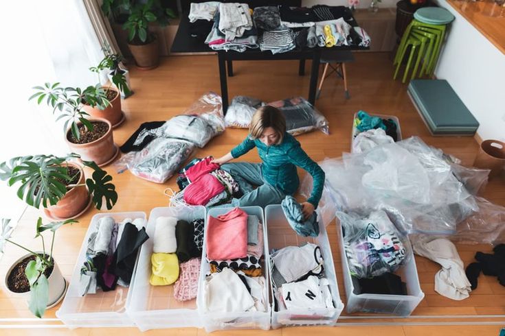 Woman organizing clothes in clear plastic bins, surrounded by folded garments and houseplants on a wooden floor.