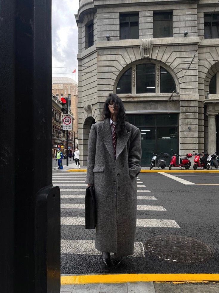 Person in a long gray coat standing on a city crosswalk near a historic building, holding a briefcase.