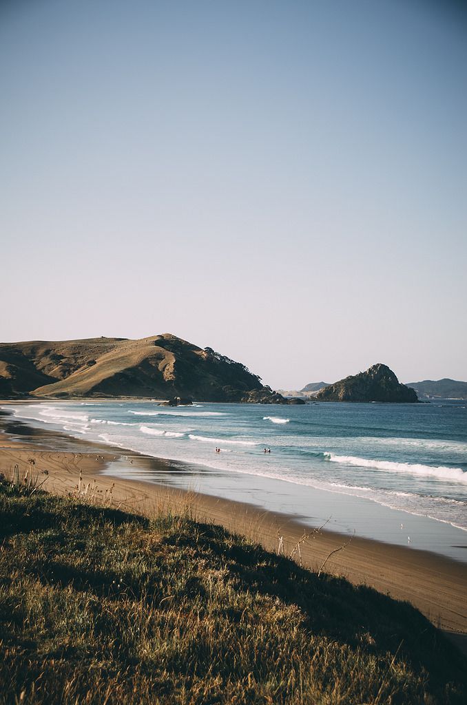 Sandy beach with gentle waves and hills in the background under a clear blue sky.
