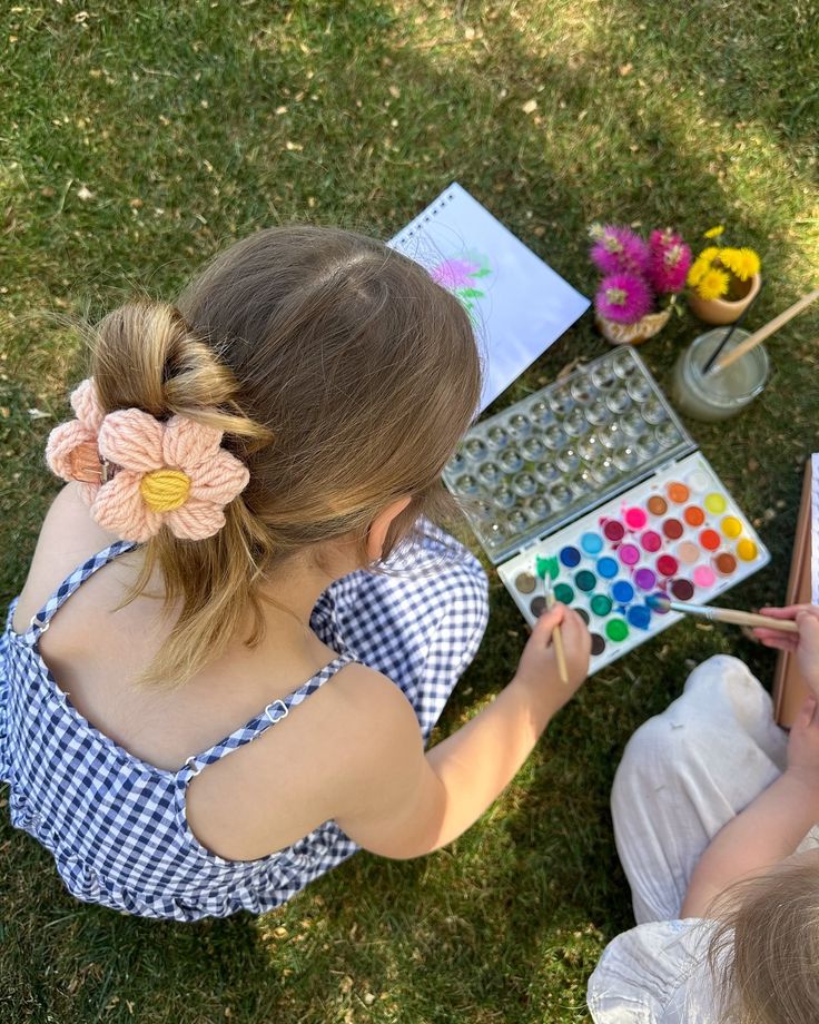 Child painting outdoors with watercolors, wearing a gingham dress and flower hair clip, on a sunny day.