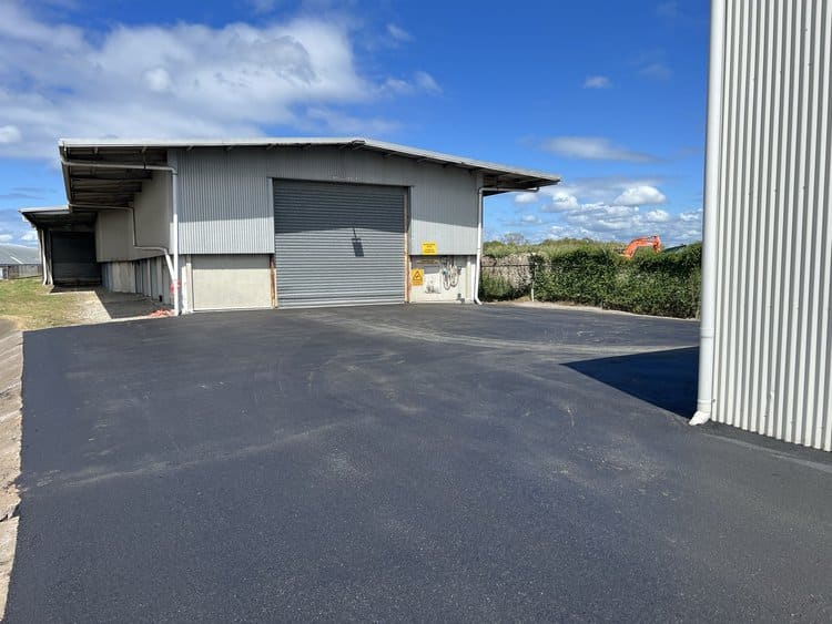 Freshly paved asphalt driveway leading to a large industrial warehouse with blue skies overhead.