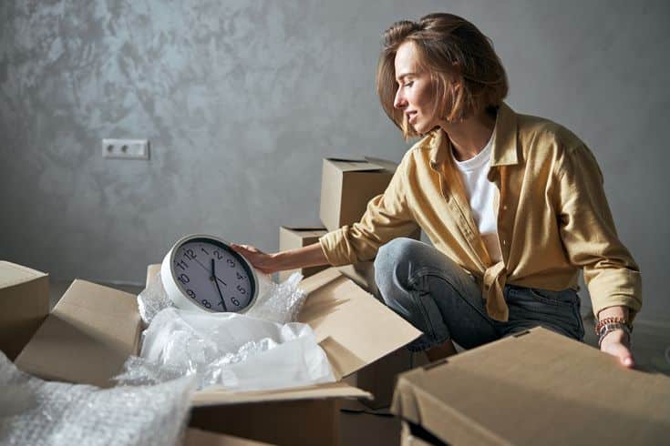 Woman unpacking clock from moving boxes in new home, surrounded by cardboard, wearing casual clothes.