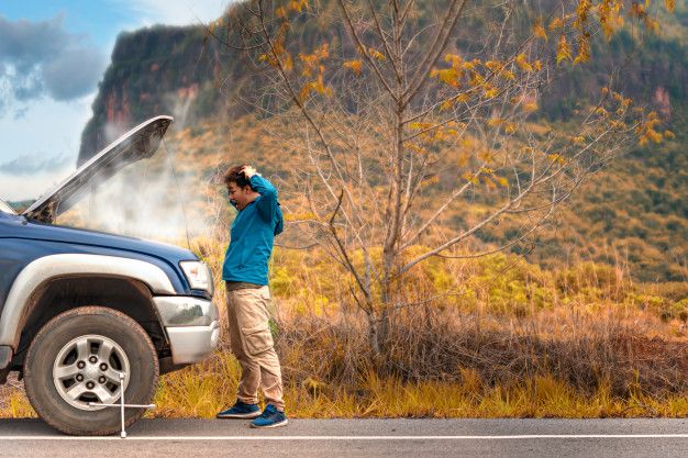 Man standing by steaming car on roadside with open hood, surrounded by autumn landscape.