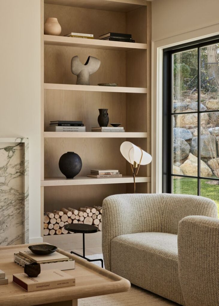 Modern living room with cozy armchair, wooden shelves, decorative vases, and books, featuring a large window view.