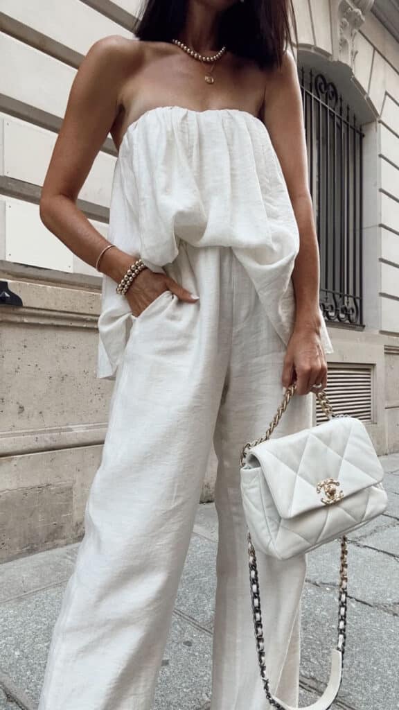 Woman in stylish white outfit with a quilted handbag, standing against a city building backdrop. Fashion street style.