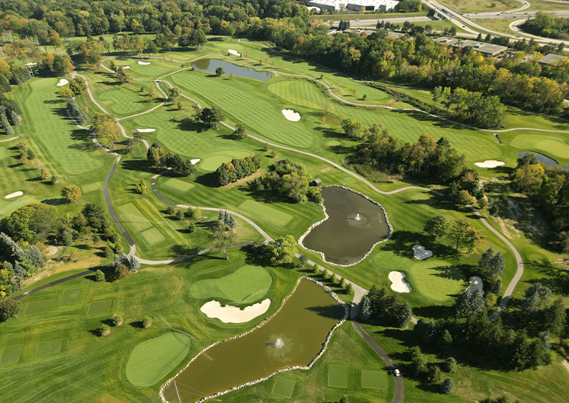Aerial view of a lush golf course with greens, ponds, sand traps, and trees on a sunny day.