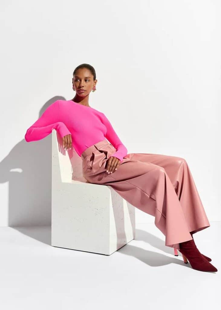 Stylish woman in pink top and pink pants sitting on modern white chair in bright studio setting.